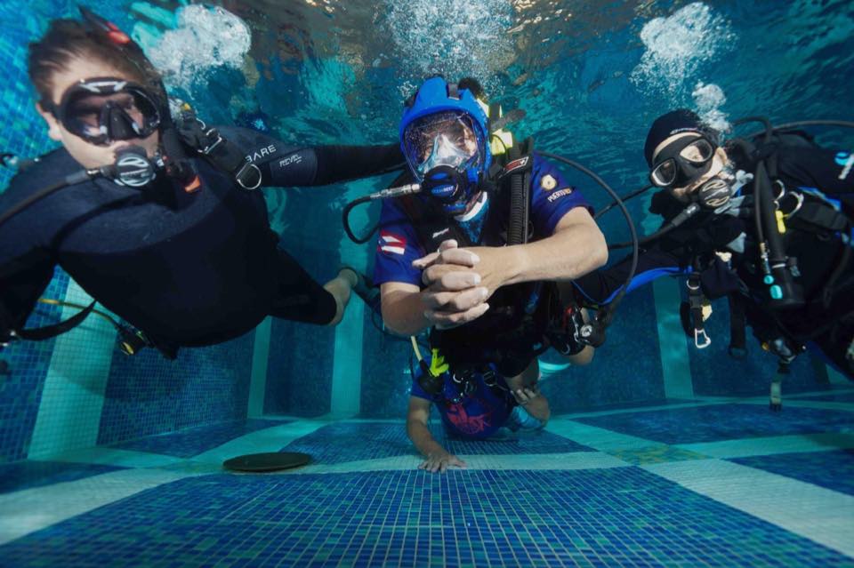 An adaptive diver in the pool with instructors on each side, everyone giving the OK