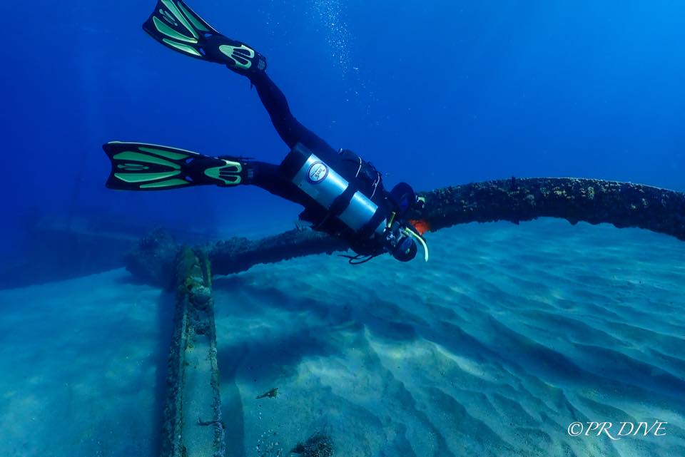 Un buzo deslizándose sobre la arena junto a una tubería vieja en agua azul profundo